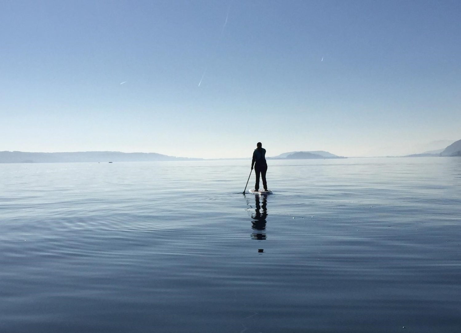Kontakt ist einfach. Und Erreichbarkeit auch. Hier sieht man einen Menschen stehend auf einem SUP StandUp Paddle bei wolkenlosem Himmel auf einem See. Im Hintergrund ein paar kleine Hügel.